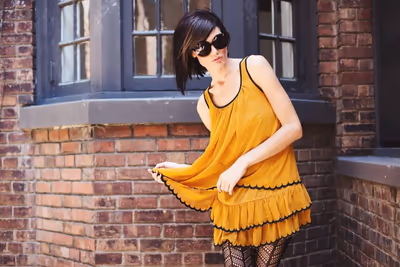 Frau in gelbem Sommerkleid mit Sonnenbrille vor einer Backsteinfassade