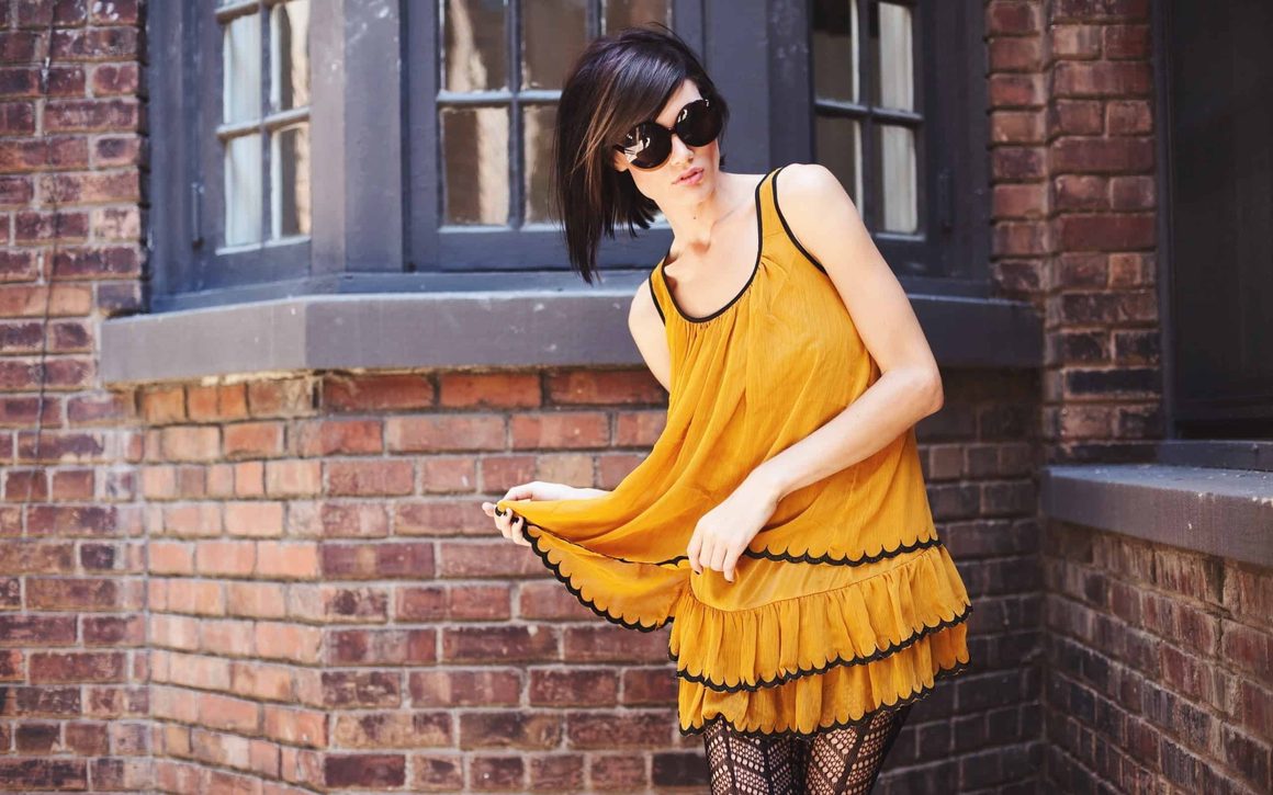 Frau in gelbem Sommerkleid mit Sonnenbrille vor einer Backsteinfassade